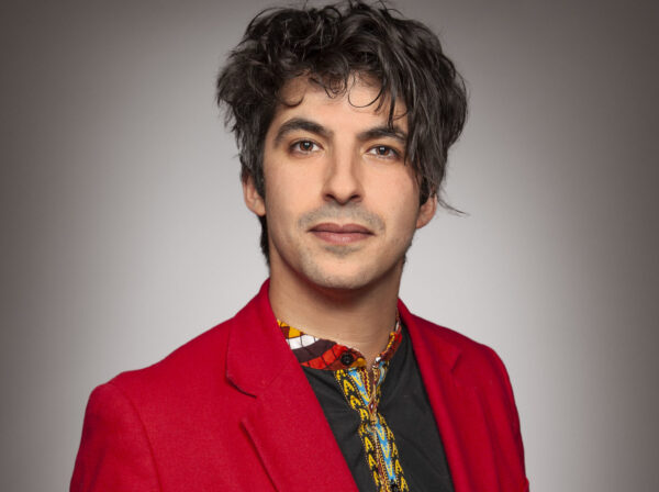 A black-haired man in a red blazer looks at the camera. / Un homme aux cheveux noirs, portant un blazer rouge, regarde la caméra.