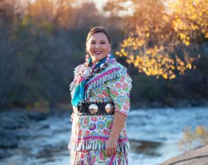 Amanda Fox is looking at the camera. Behind her we can see a river and a forest with yellow leaves.